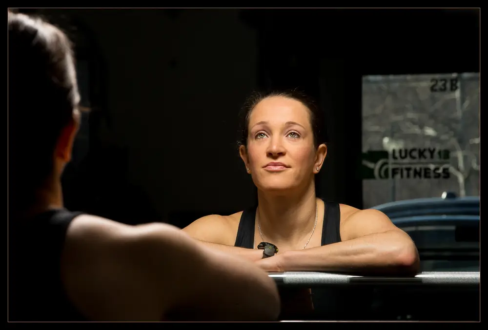 Lucky13Fitness-8081MD_LowResDL A woman in a black tank top with a focused expression looks up, resting her arms on a barbell in a gym.